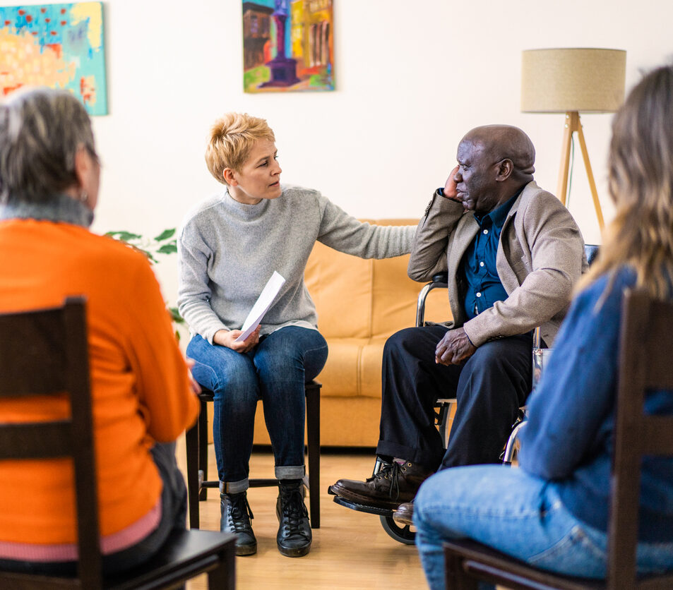 Placing her hand onto a man's shoulder, a healthcare professional is giving comfort to a senior man in a wheelchair, during a support meeting in a nursing home, helping the elderly residents