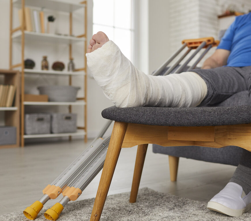 Senior man with broken leg in gypsum plaster cast sitting on sofa at home and talking on mobile phone. Elderly patient with injured foot resting on couch and calling doctor on cellphone. Close up
