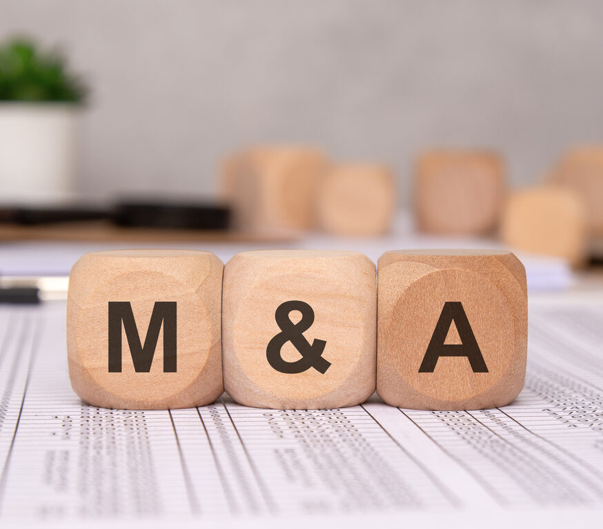 Three wooden cubes on financial spreadsheets, representing business mergers and acquisitions. blurred office background, with documents and a laptop, emphasizes corporate finance, investment strategy