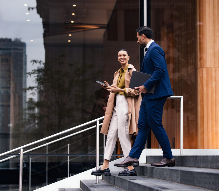 Two business professionals, a woman and a man, walk down exterior stairs beside a glass-fronted building. They converse and carry a tablet and folders, projecting a confident, city-work vibe.