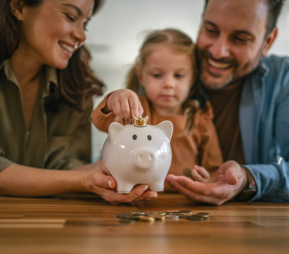 Young family, consisting of parents and their daughter, engaging in financial education, with the child putting coins into a white piggy bank and parents assisting with the saving process